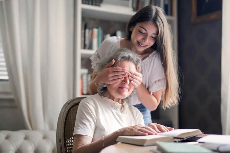 happy granddaughter playing guess who game with aged grandmother while making surprise in light living room of modern apartment